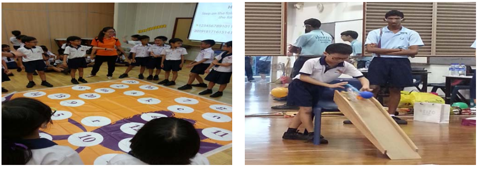 Students in uniforms play on a numbered floor mat, while another student rolls a ball down a wooden ramp indoors.
